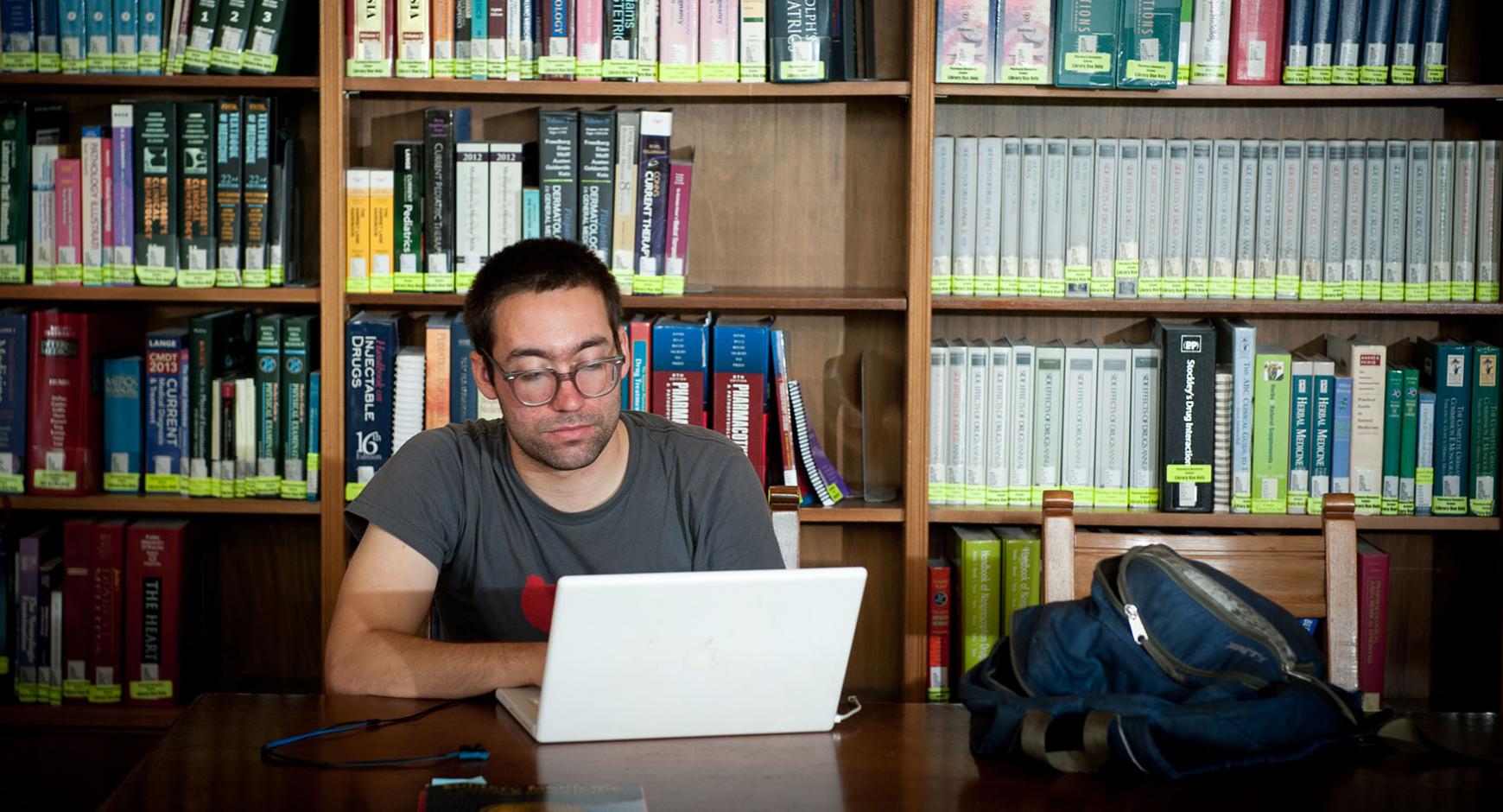 man in glasses works on laptop in front of bookcases in classic library space