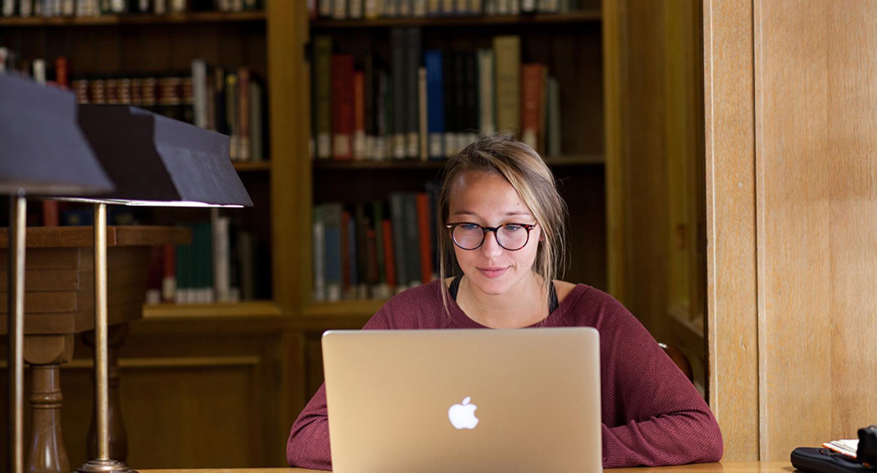 college student in glasses smiles while viewing laptop screen, books in background